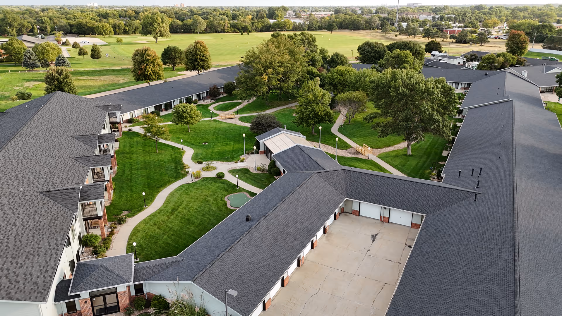 Aerial view of Riverside Lodge Retirement Community showing multiple connected buildings with gray roofs surrounding a landscaped courtyard with green lawns, trees, walking paths, and outdoor lighting. Beyond the community are open fields and more trees under a clear sky.