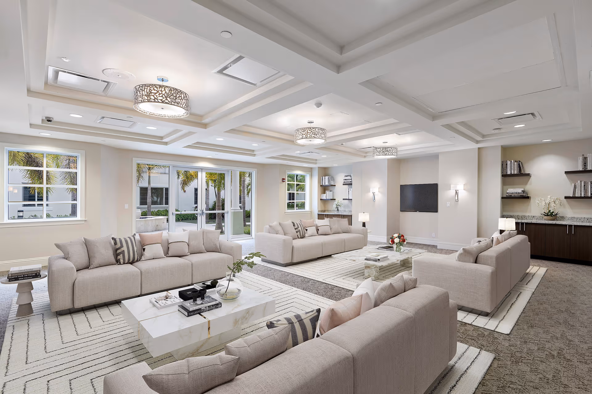 A spacious and bright senior living facility lounge with multiple beige sofas arranged around white marble coffee tables. The room features large windows and glass doors letting in natural light, modern ceiling lights, built-in shelves with books and decor, a wall-mounted flat-screen TV, and a carpeted floor with area rugs.