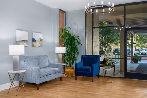 A modern living room area with a light blue loveseat, a dark blue armchair, two side tables each with a lamp, a large potted plant, and two abstract paintings on a light gray wall. The room has wooden flooring and large glass doors showing an outdoor view with trees and parked cars.