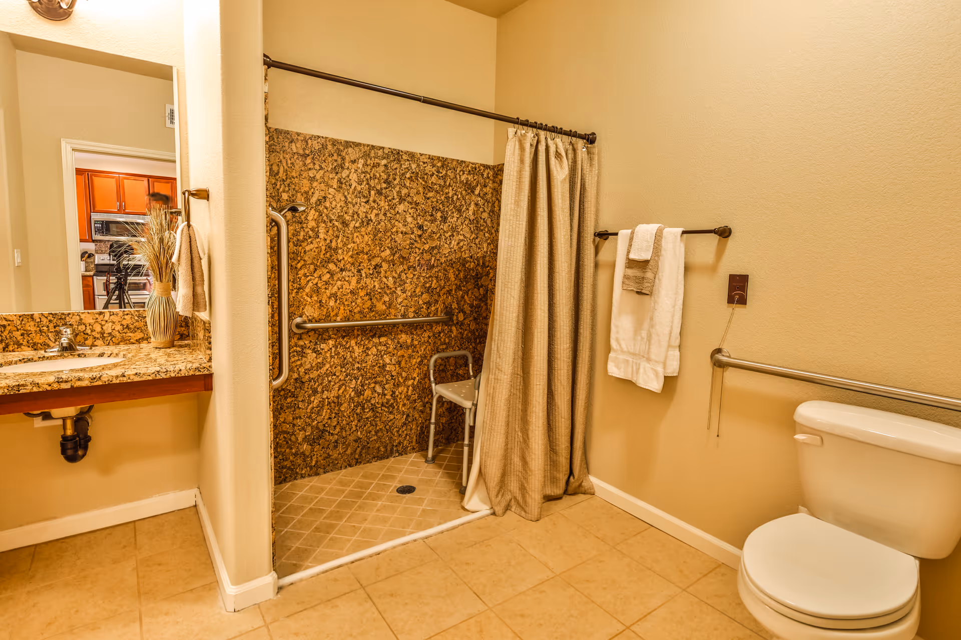 A bathroom with a walk-in shower featuring a granite wall and a shower chair. There is a toilet with grab bars on the wall, a towel rack with white towels, and a sink with a granite countertop and a decorative vase with dried plants. The bathroom has beige tiled floors and walls.