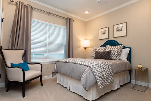 A neatly made bed with gray and white patterned bedding and multiple pillows in a bedroom. Next to the bed is a small side table with a flower vase and a floor lamp. A beige armchair with a blue cushion is placed near a window with closed blinds and gray curtains. Two framed pictures hang on the beige wall above the bed.