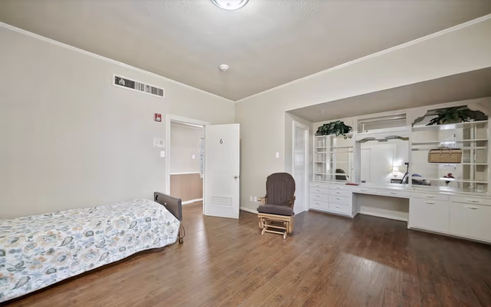 A simple bedroom with a single bed covered with a floral bedspread on the left, a wooden rocking chair with a cushion in the center, and a built-in white vanity with shelves, drawers, and plants on the right. The room has wooden flooring, beige walls, and a door marked with the number 6 that is slightly open.