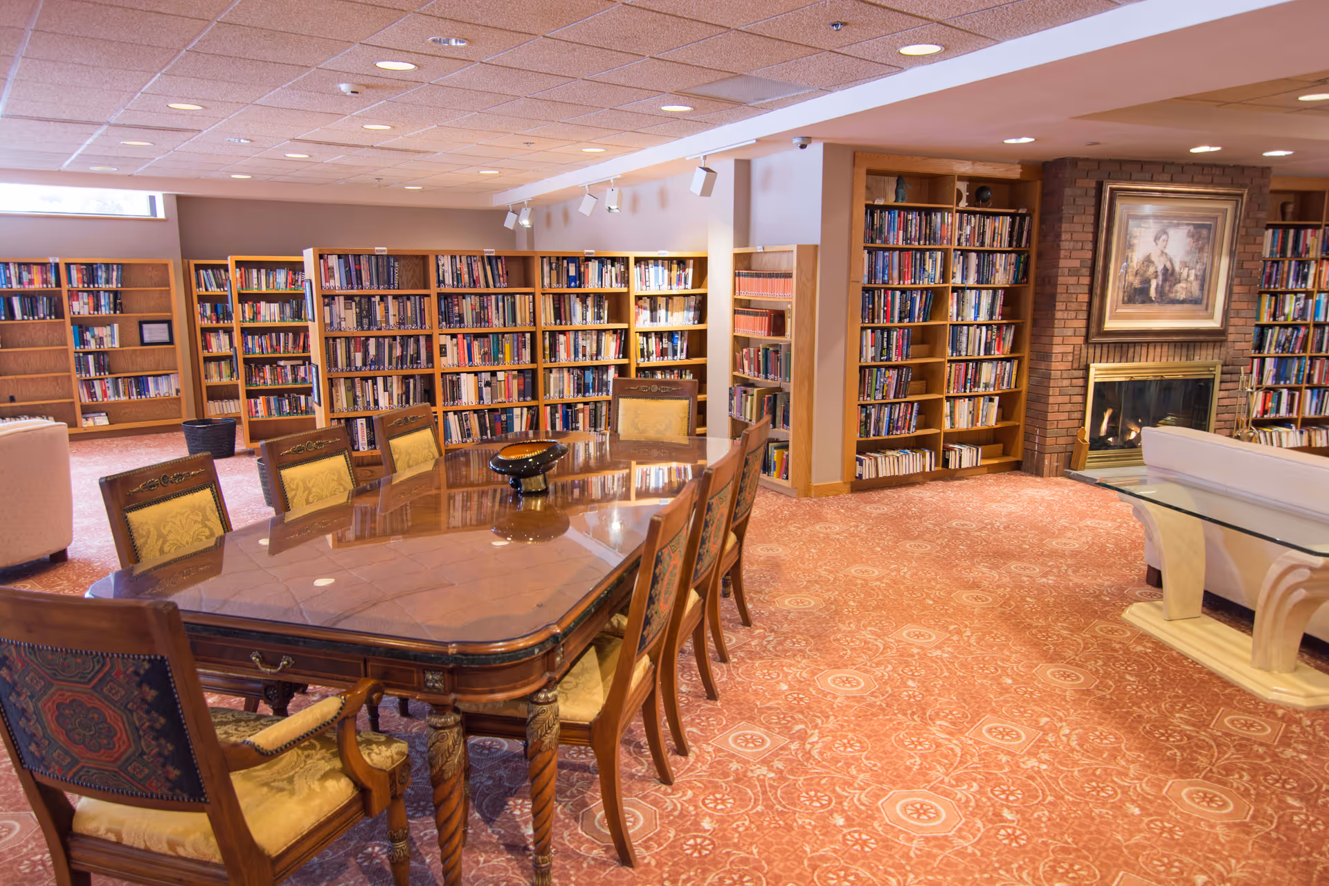 A spacious library-style common room with a large wooden table and chairs, bookshelves lining the walls, and a fireplace.