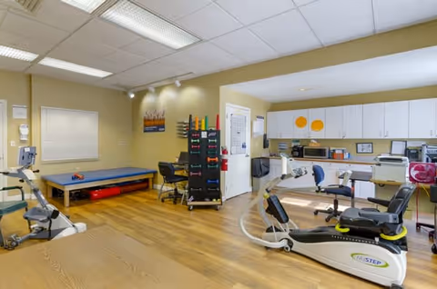 A physical therapy or rehabilitation room with exercise equipment including stationary bikes and a treatment table. There are chairs, a rack with colorful exercise bands, cabinets, and a countertop with various supplies. The room has wood flooring and fluorescent ceiling lights.