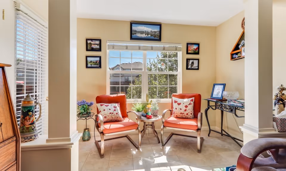 A bright sitting area with two orange cushioned chairs featuring floral pillows, positioned around a small round table with decorative plants. The space is illuminated by natural light from a large window with a view of trees and houses outside. The walls are adorned with framed pictures, and there is a small side table with additional decor items.