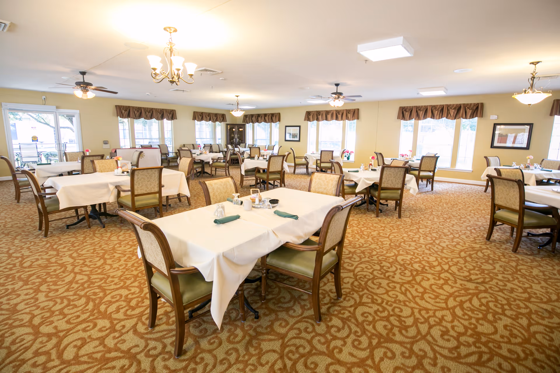 Dining room with several tables covered in white tablecloths, wooden chairs, chandeliers, and large windows.