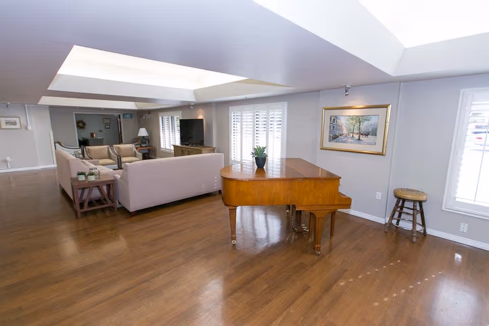 A spacious senior living facility common area with wooden flooring, a grand piano with a small plant on top, a beige sectional sofa set with a side table holding small plants, a flat-screen TV on a wooden stand, large windows with white shutters, a framed painting on the wall, and a wooden stool near the window.
