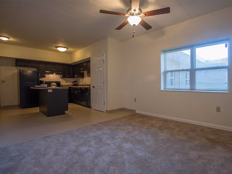 Interior view of a senior living facility apartment at Morton Estates showing a combined living room and kitchen area. The living room has carpeted flooring and a ceiling fan with a light fixture. The kitchen features dark cabinetry, a black refrigerator, stove, dishwasher, and a small island. There is a window with blinds on the right wall allowing natural light into the room.