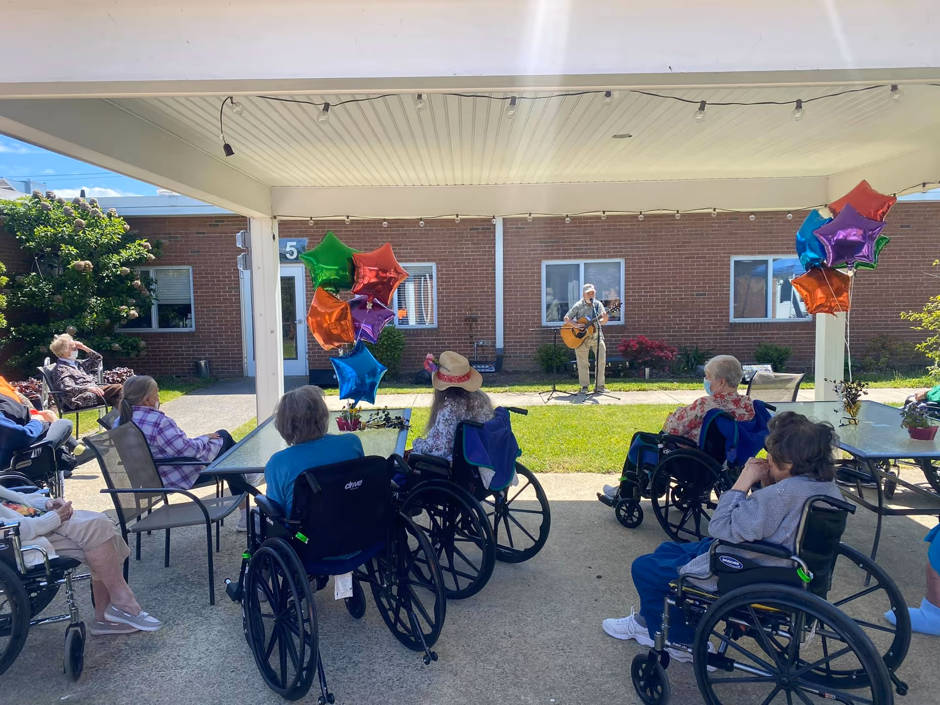 A group of elderly people, many in wheelchairs, seated outdoors under a covered patio watching a man playing guitar and singing on a grassy area in front of a brick building. Colorful star-shaped balloons are tied to the patio posts.