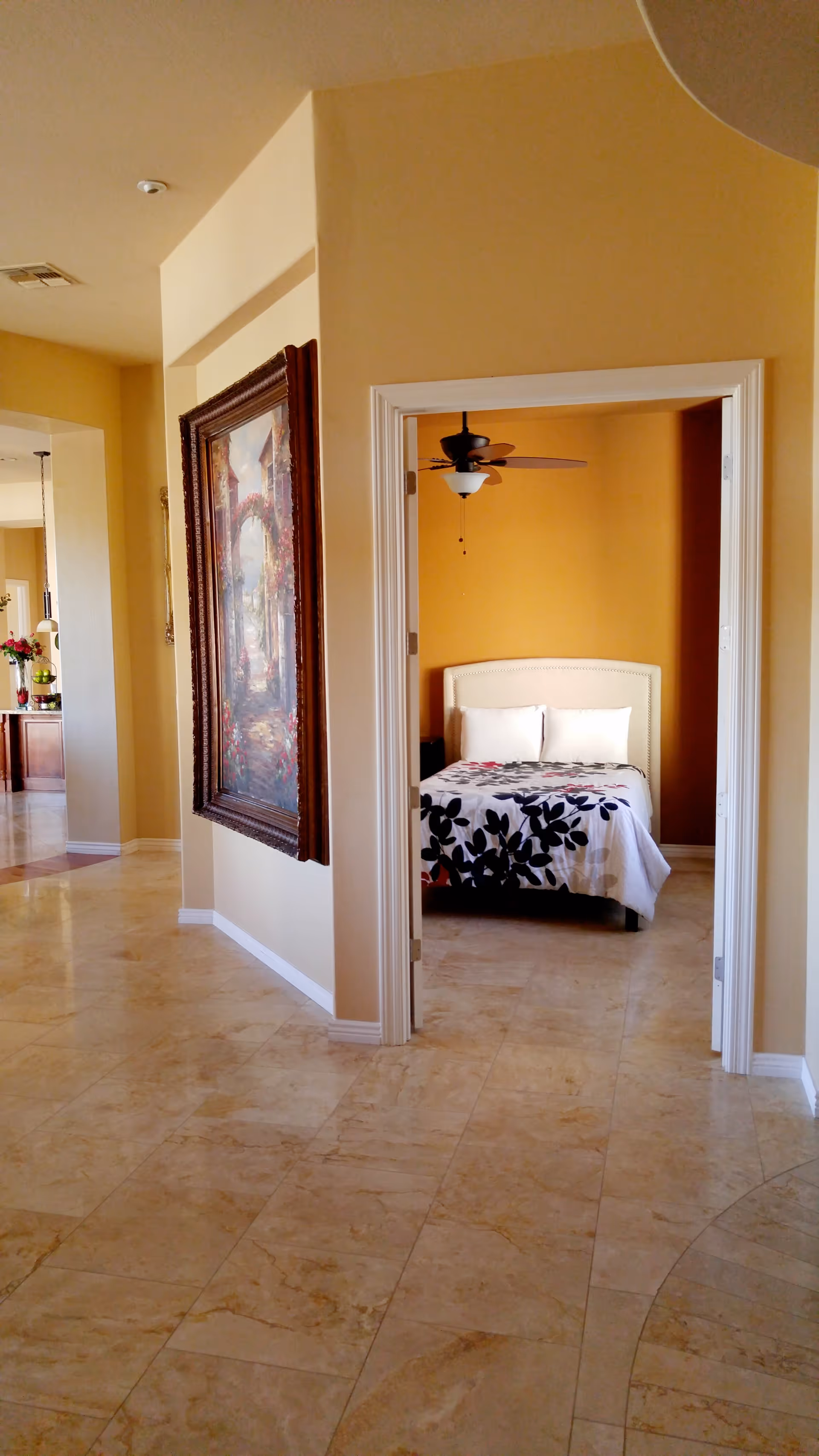 A tiled hallway looking into a bedroom with a bed, ceiling fan, and a large framed painting on the left wall.