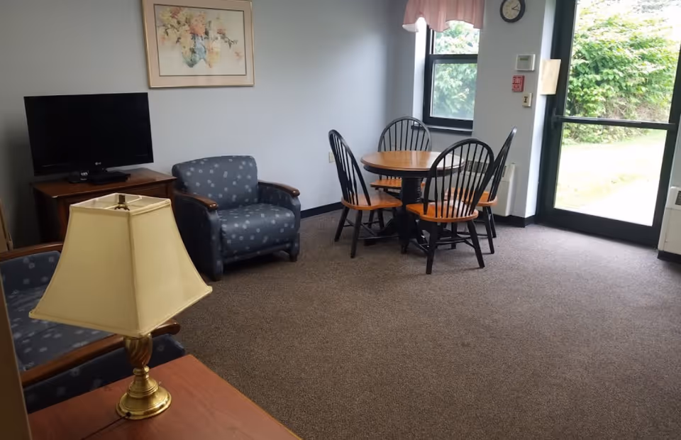 A cozy room with a small round wooden dining table and four chairs near a window and glass door. There are two blue armchairs with a subtle pattern, a wooden TV stand with a flat-screen TV, a framed floral painting on the wall, and a table lamp with a beige shade on a wooden side table. The room has carpeted flooring and light-colored walls.