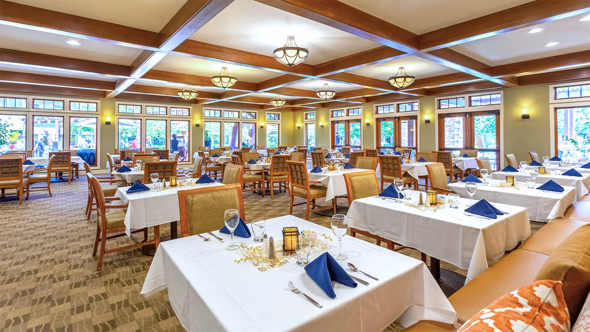 A spacious dining room with multiple tables covered in white tablecloths, each set with blue napkins, glassware, and silverware. The room features large windows allowing natural light to fill the space, wooden ceiling beams, and warm lighting fixtures. Chairs are arranged neatly around the tables, and there is a cushioned bench with decorative pillows along one side.
