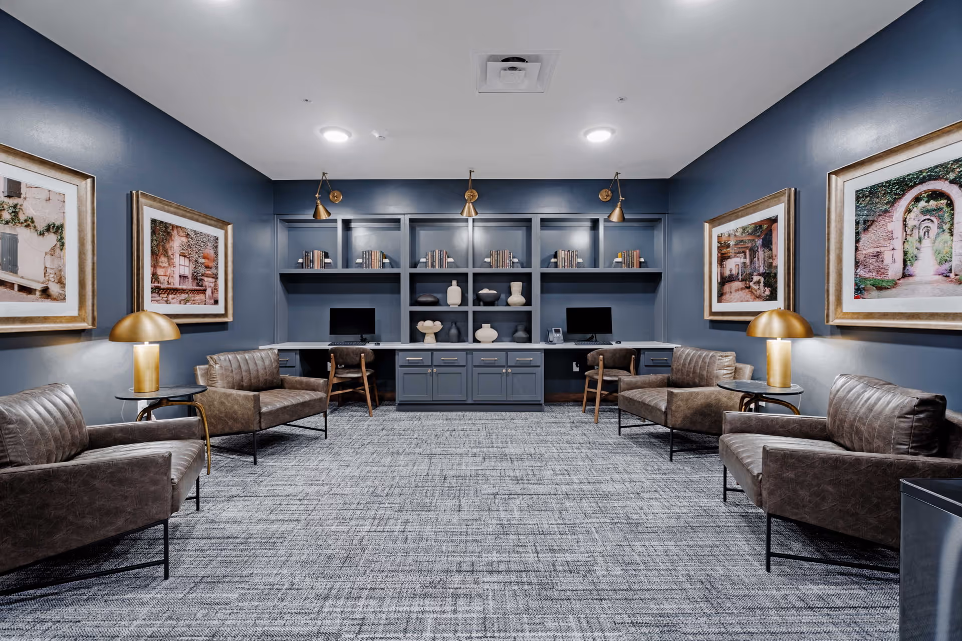 A modern senior living common area with four brown leather armchairs arranged around two gold table lamps on small side tables. The walls are painted blue and decorated with framed pictures of outdoor scenes. At the far end of the room, there is a built-in shelving unit with books, decorative vases, and two computer workstations with chairs. The floor is covered with a gray patterned carpet.