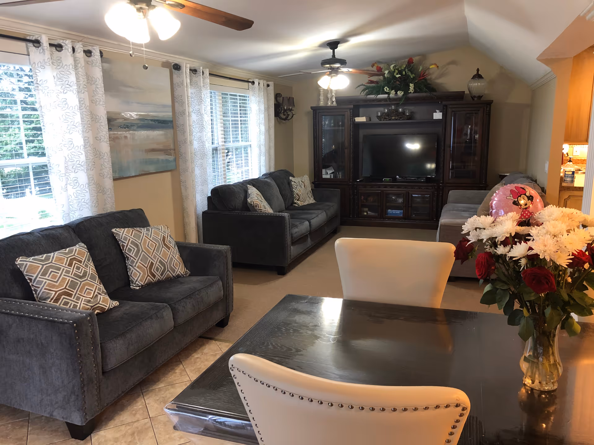 A cozy living room with two dark gray sofas adorned with patterned cushions, a large wooden entertainment center with a flat-screen TV, and a ceiling fan with lights. The room has large windows with white patterned curtains letting in natural light. In the foreground, there is a dark wooden dining table with cream-colored chairs and a vase of red and white flowers. A Minnie Mouse balloon is visible on the right sofa near the kitchen entrance.