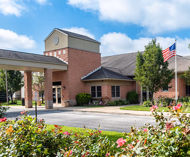 Front entrance of a brick senior living facility with a covered drive, landscaped flowers, and an American flag.