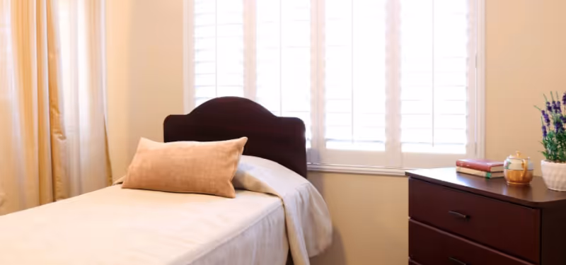 A neatly made single bed with a wooden headboard beside a window with shutters and a dresser topped with books and a potted plant.