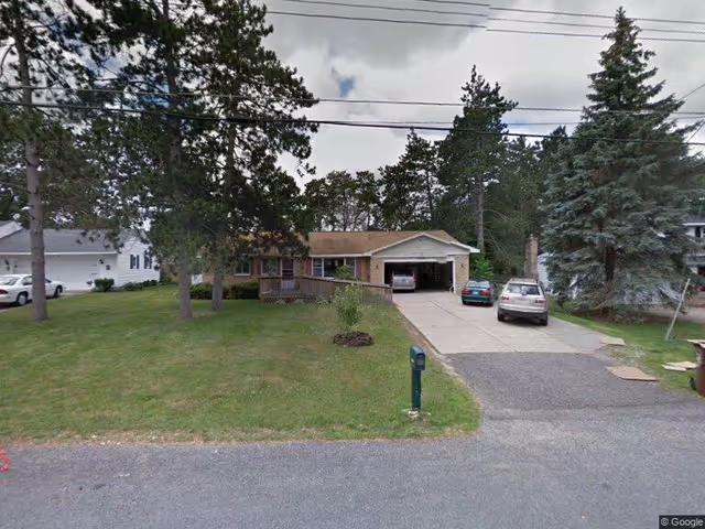 Single-story residential house with a brown roof and attached garage, surrounded by green grass and tall trees. There are three cars parked in the driveway and a mailbox near the street. The sky is partly cloudy.