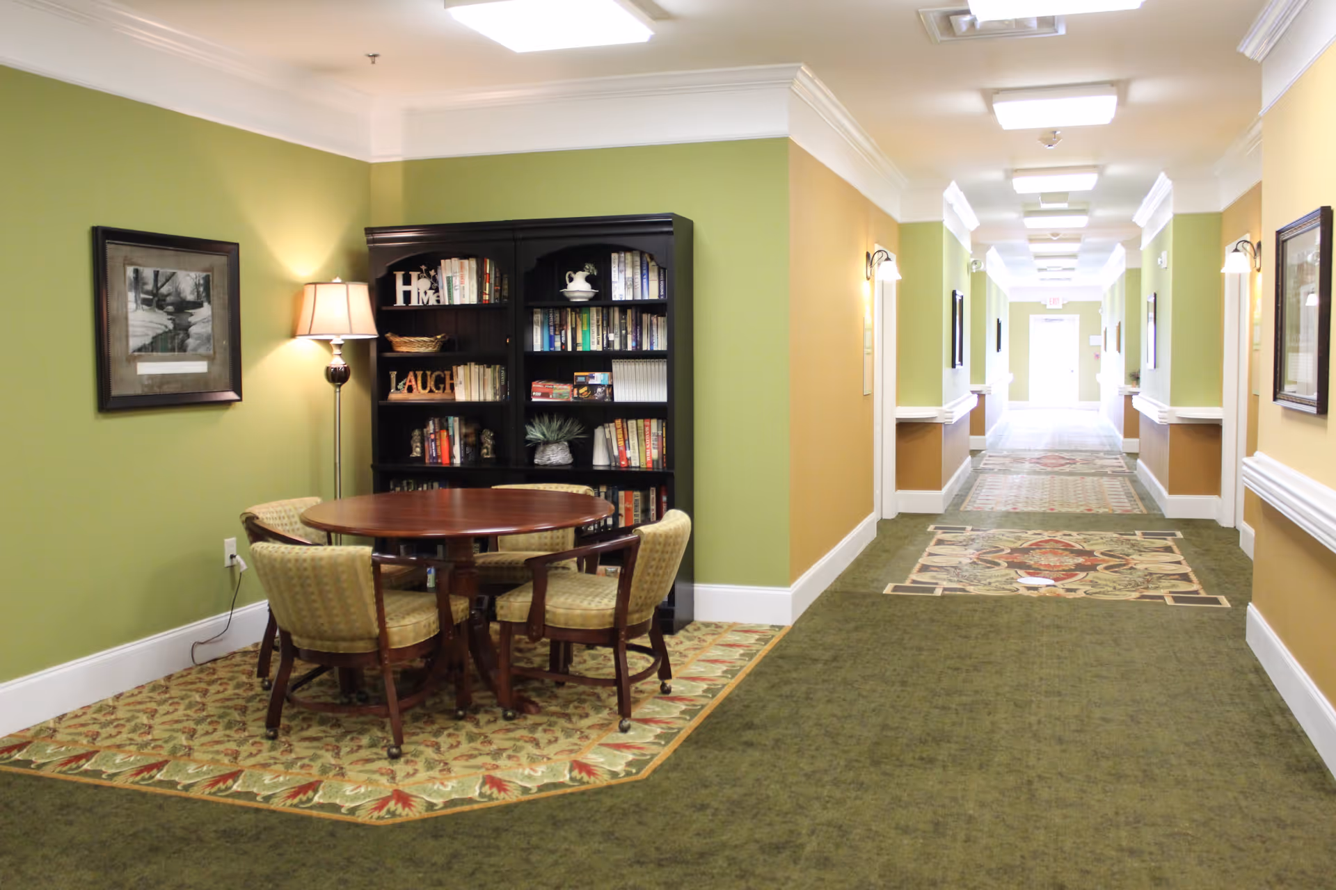 A hallway in a senior living facility with green and yellow walls, white crown molding, and patterned green carpet. On the left side, there is a small seating area with a round wooden table, four upholstered chairs, a floor lamp, a black bookshelf filled with books and decorative items, and a framed picture on the wall.