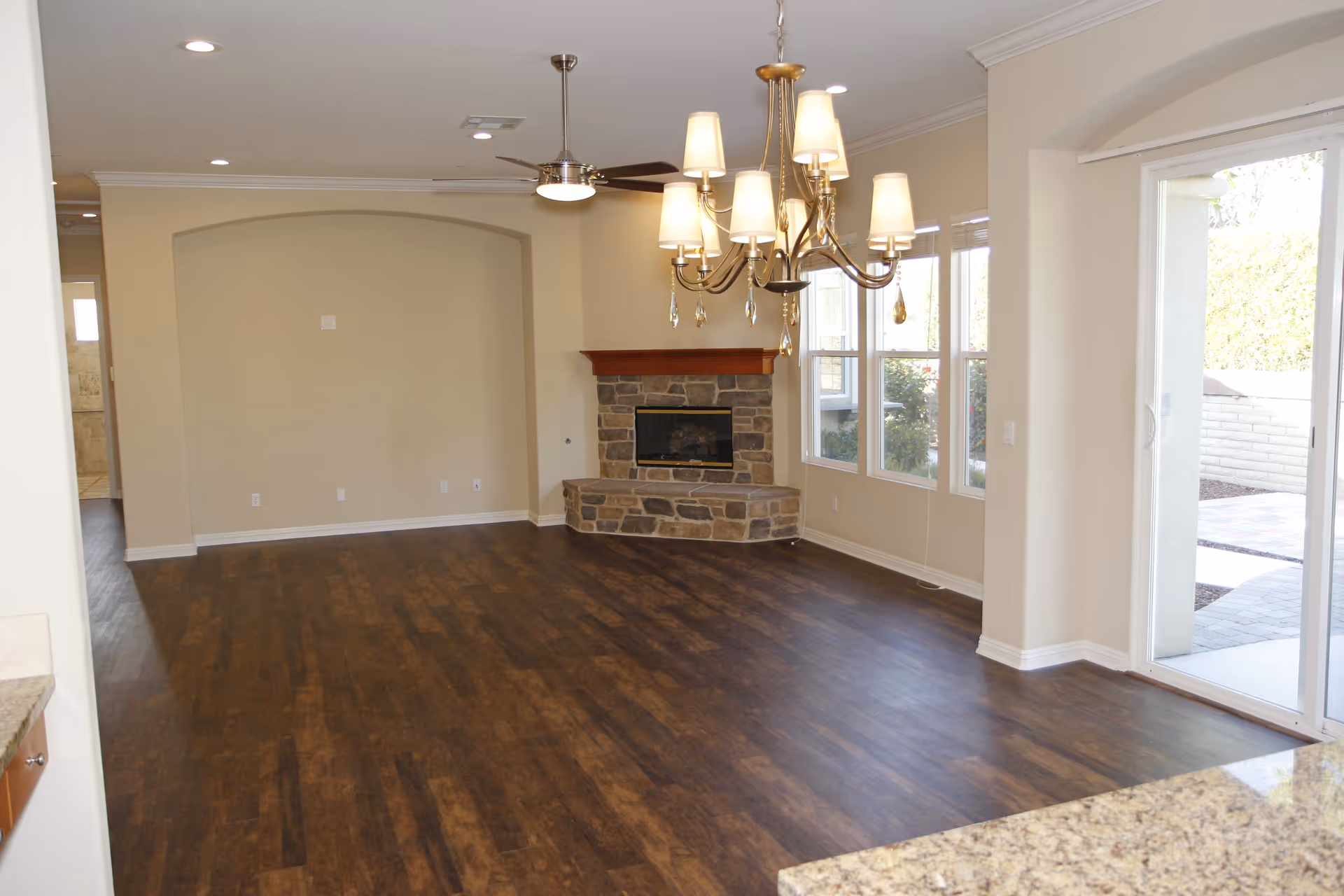Empty living room with dark wood flooring, a stone fireplace with a wooden mantle, a ceiling fan with a light, a chandelier with multiple lampshades, large windows letting in natural light, and a sliding glass door leading to an outdoor patio area.