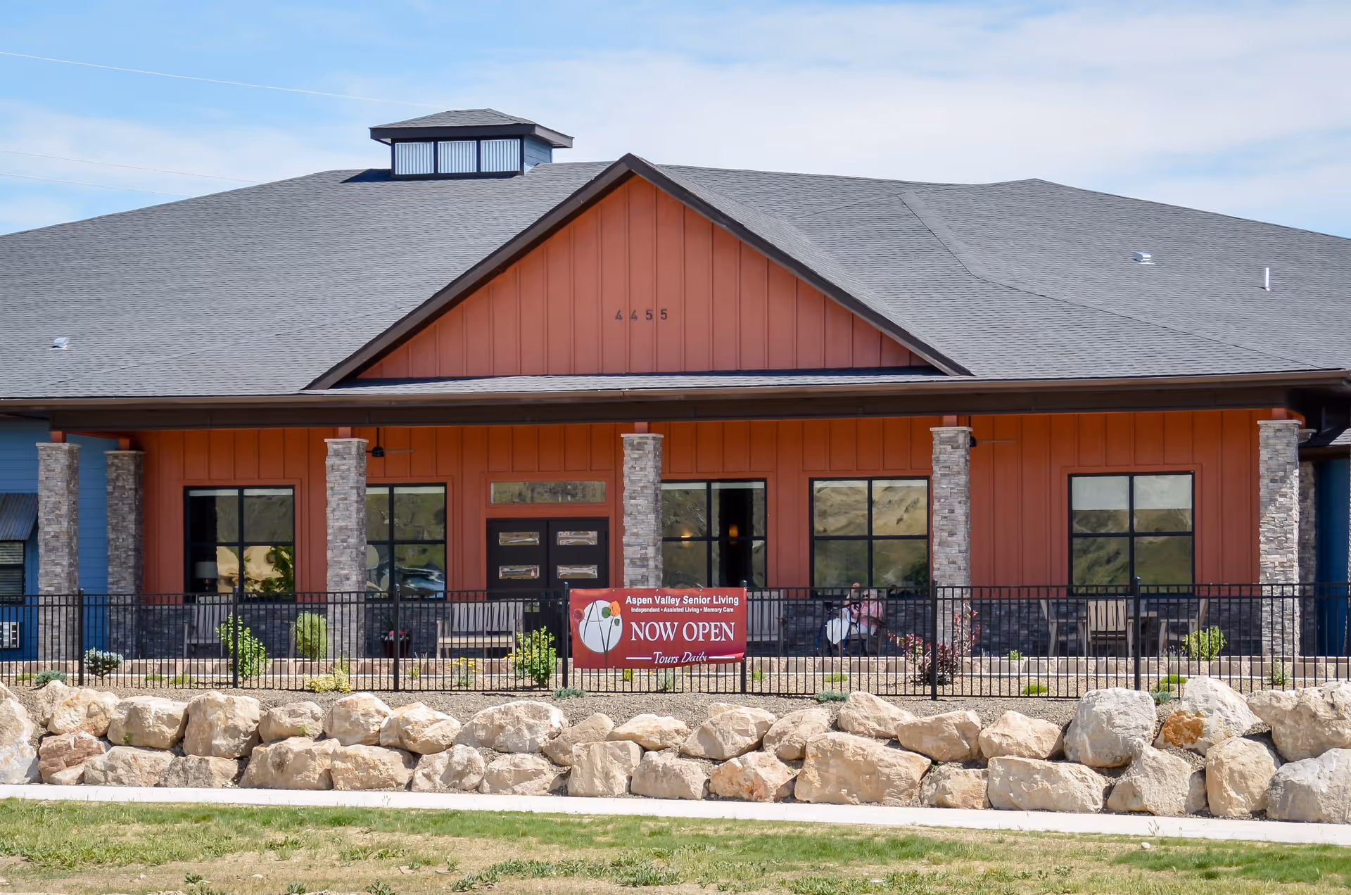 Front exterior view of Aspen Valley Senior Living building with a stone and wood facade, large windows, and a sign that reads 'Aspen Valley Senior Living NOW OPEN Tours Daily' in front of a black metal fence and rock landscaping.