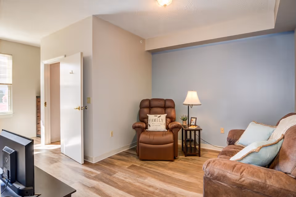 A cozy living room area with a brown leather recliner chair featuring a decorative pillow that says 'FAMILY', a small side table with a lamp, a small plant, and a framed photo. There is also a brown couch with light blue and white pillows. The walls are painted beige and light blue, and the floor has wood-style flooring. A TV is visible on a stand in the foreground, and an open door leads to another room with a window.