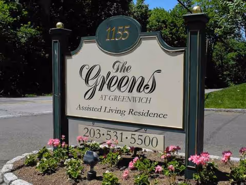 A large sign for The Greens at Greenwich Assisted Living Residence with pink flowers nearby.