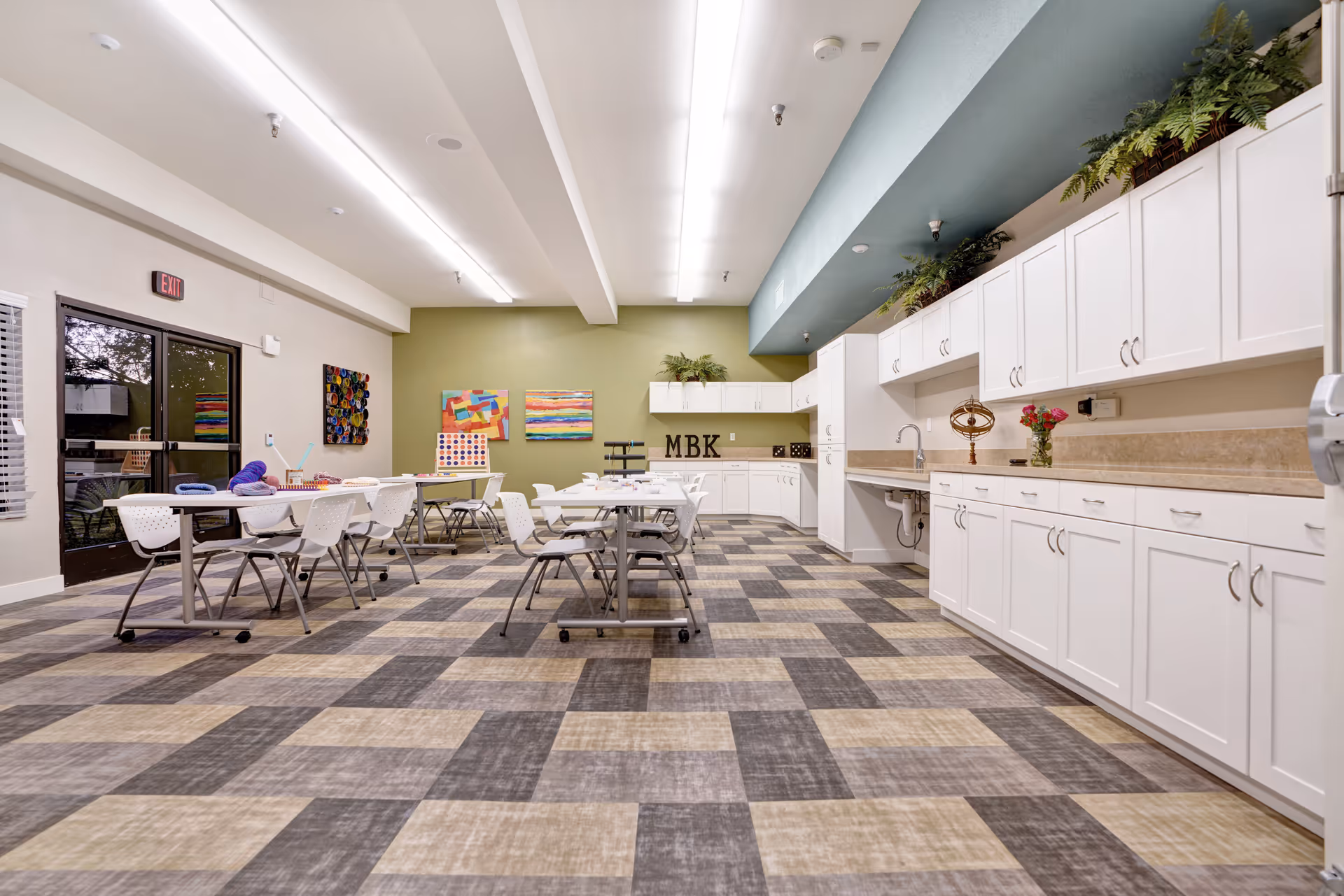 A spacious, well-lit activity room with checkered flooring, white cabinets along one wall, and several tables and chairs arranged for group activities. Colorful artwork is displayed on a green accent wall, and plants are placed on top of the cabinets. There is a door with windows on the left side leading outside.