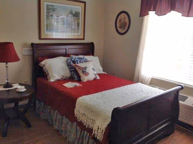 A cozy bedroom with a dark wooden bed frame, red bedspread, white throw blanket, and decorative pillows. There is a round wooden side table with a red lamp and a cup and saucer. The room has a window with white curtains and a maroon valance, a framed picture above the bed, and a small round floral wall decoration.