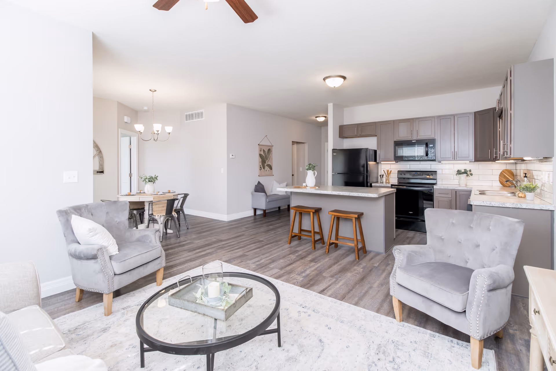 A bright and modern open-concept living space featuring a kitchen with gray cabinets, black appliances, and a kitchen island with two wooden stools. The living area includes two gray upholstered armchairs and a round glass coffee table on a light-colored rug. In the background, there is a dining area with a wooden table and metal chairs under a chandelier, and a small sitting area with a gray armchair against a light gray wall.