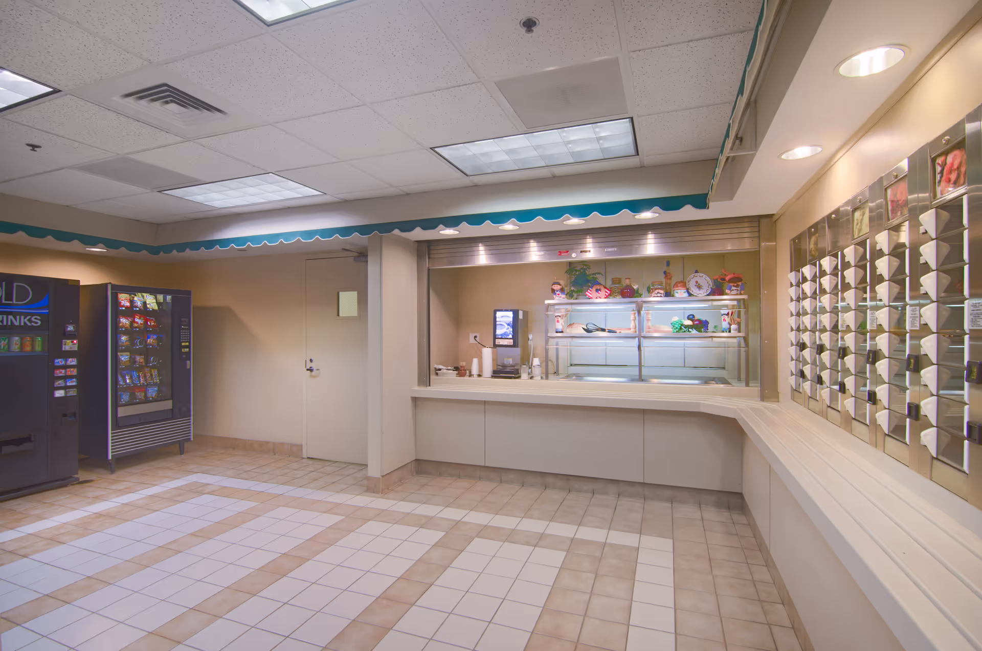 Interior view of a senior living facility's snack and drink area with vending machines on the left, a counter with a beverage dispenser in the center, and a wall-mounted mail or key organizer on the right. The floor is tiled and the ceiling has fluorescent lighting.