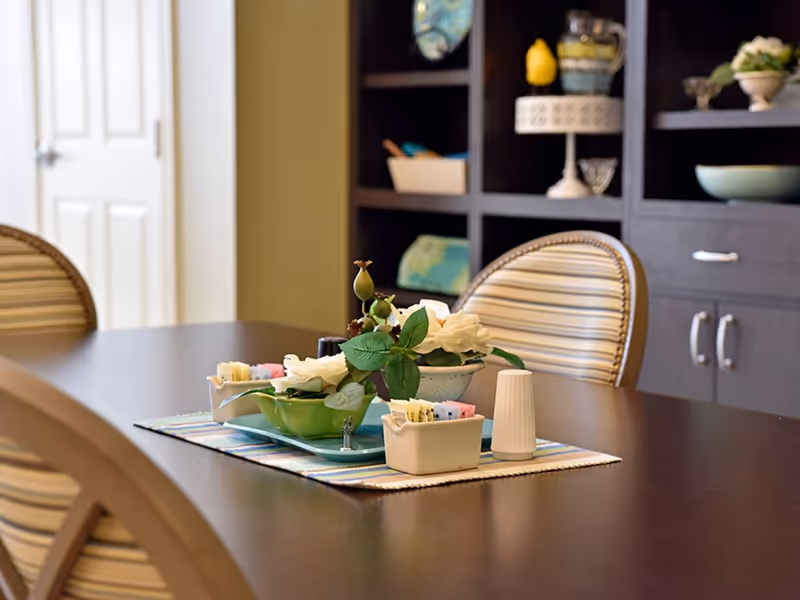 A dining table with a striped table runner, decorated with a small floral arrangement, a container holding sugar packets, and a salt shaker. The table is surrounded by chairs with striped upholstery. In the background, there is a dark wooden shelving unit with various decorative items and a closed white door.