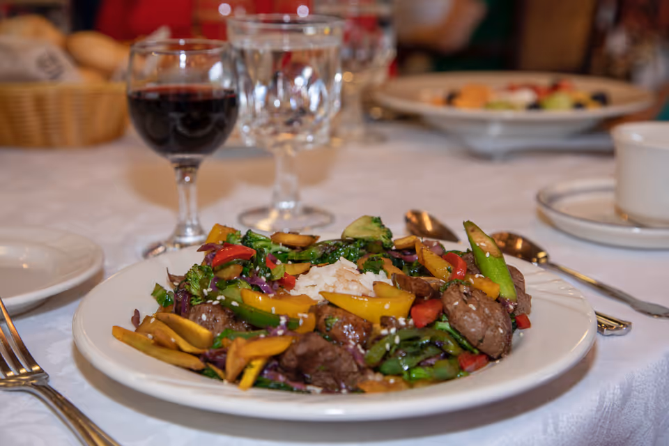 A plate of stir-fried vegetables and beef with a serving of white rice in the center, placed on a white tablecloth. In the background, there is a glass of red wine, empty wine glasses, a basket of bread, and another plate with food.