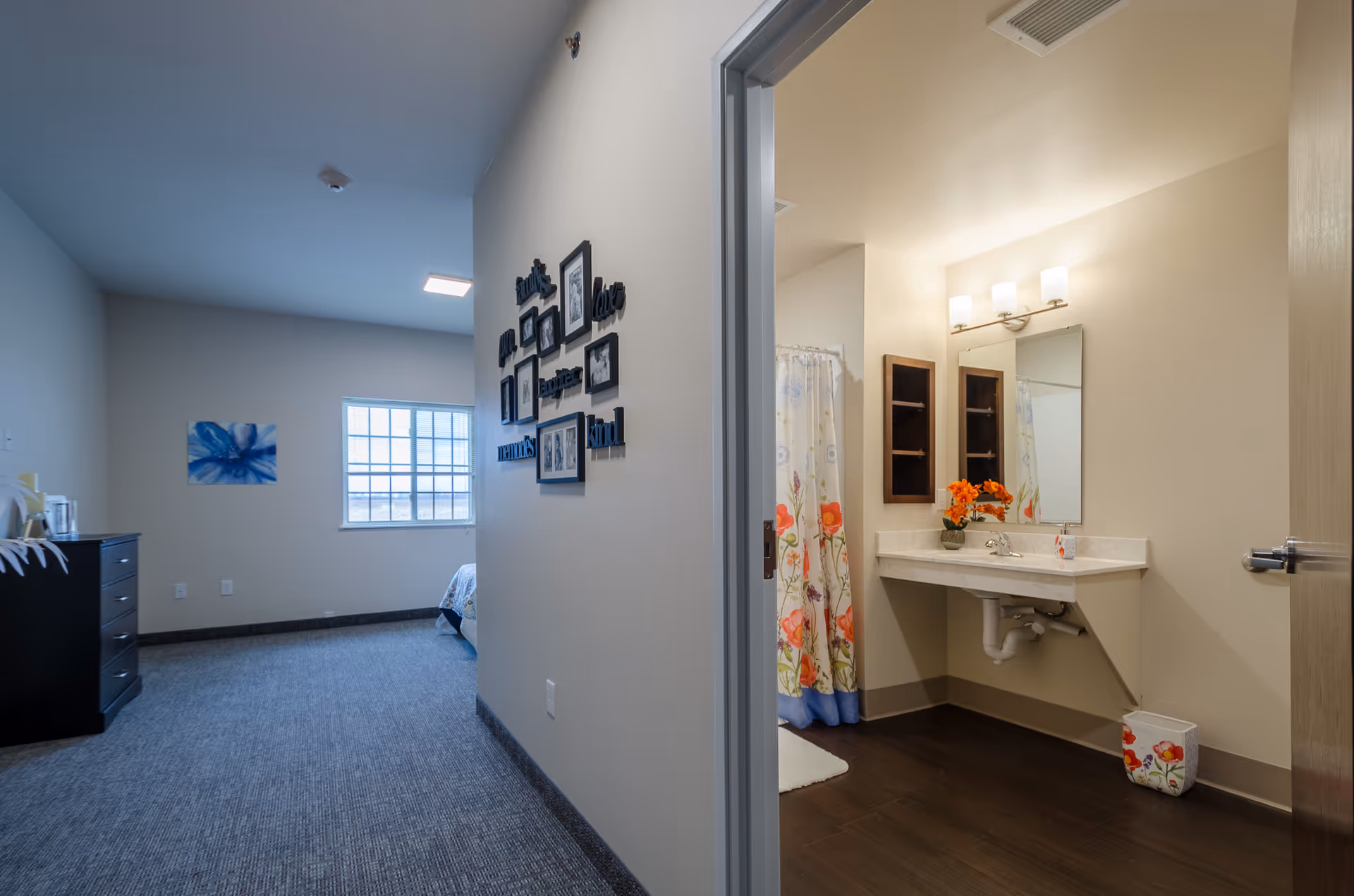 View of a senior living facility room showing a bedroom area with a window, dresser, and wall decor, and an adjacent bathroom with a sink, mirror, floral shower curtain, and decorative flowers on the counter.