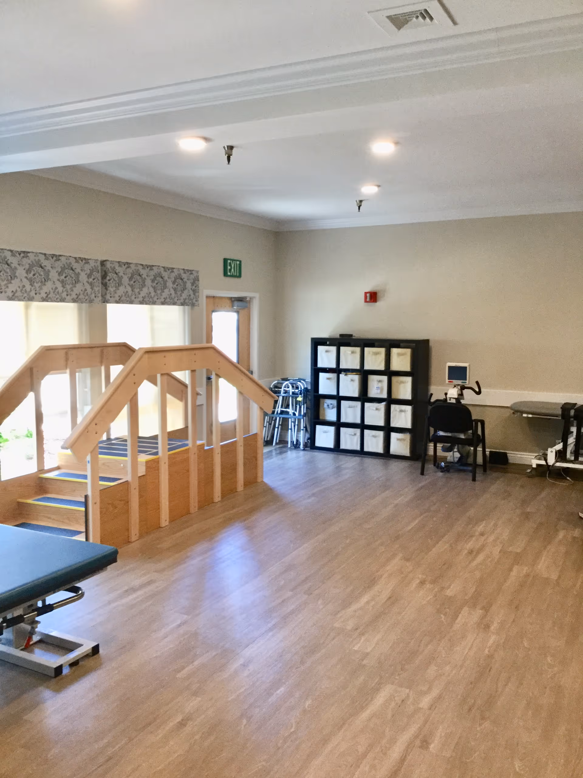 A spacious room with wooden flooring featuring a small wooden staircase with handrails, a therapy table, a black shelving unit with white storage bins, a chair with a small monitor on a stand, and several walkers near an exit door with an illuminated green exit sign.