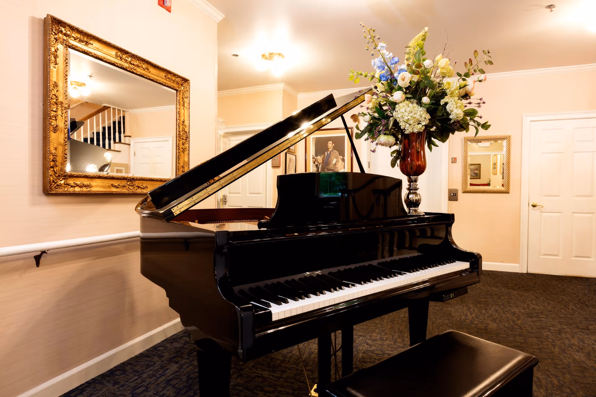 A black grand piano with its lid open is placed in a warmly lit room. On top of the piano is a large floral arrangement in a tall vase. The room features beige walls, a dark carpeted floor, a large ornate gold-framed mirror on the left wall, and a smaller mirror on the right wall. There are white doors and framed artwork visible in the background.