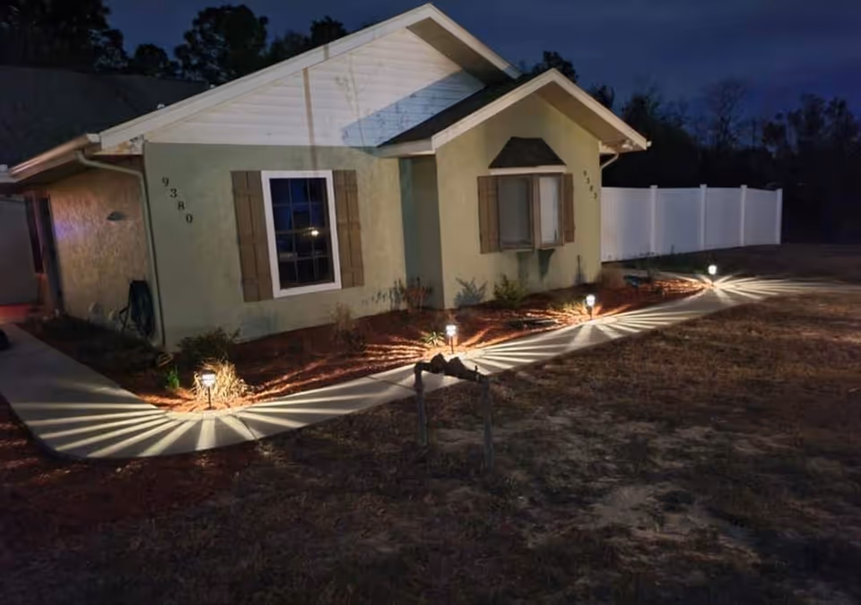 Night view of a single-story house with exterior lights illuminating a curved concrete pathway and landscaping. The house has light green walls, two windows with brown shutters, and a white fence in the background.