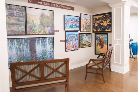 An art gallery area inside The Wellington at Lake Manassas featuring several framed paintings on a white wall. The paintings depict various scenes including landscapes and people. There are two wooden chairs with cushioned seats positioned near the wall. The floor is wooden, and a sign above the paintings reads 'Art Gallery at The Wellington.'