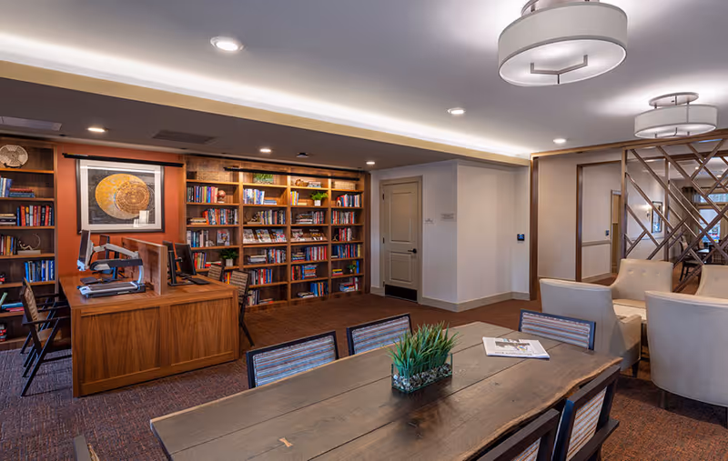 A cozy senior living facility common area featuring a large wooden table with chairs, a small plant centerpiece, and a newspaper. In the background, there is a wooden bookshelf filled with books and decorative items, a desk with computer monitors, and comfortable armchairs near a decorative partition. The room is well-lit with ceiling lights and has a warm, inviting atmosphere.