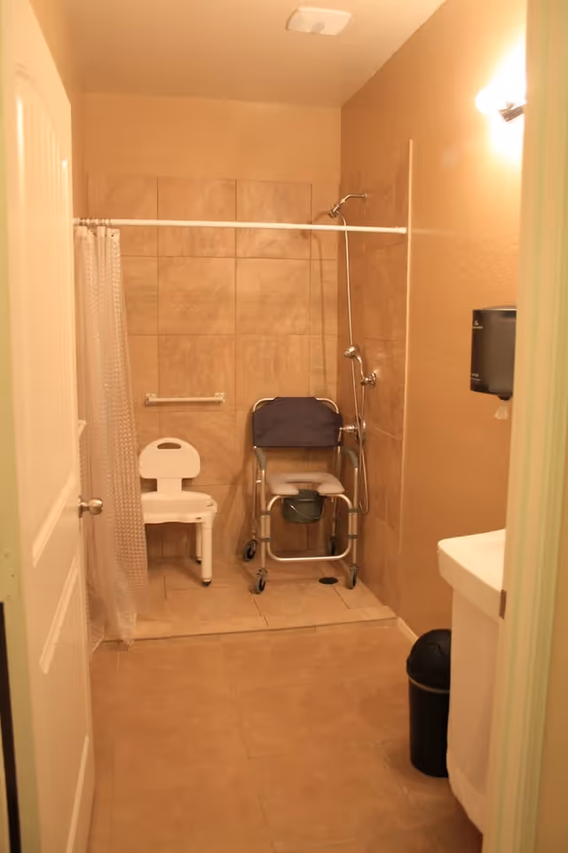 A bathroom with beige tiled walls and floor, featuring a shower area with a white shower curtain, a white plastic shower chair, and a wheeled commode chair. There is a wall-mounted soap or paper dispenser and a small trash can next to a white sink.