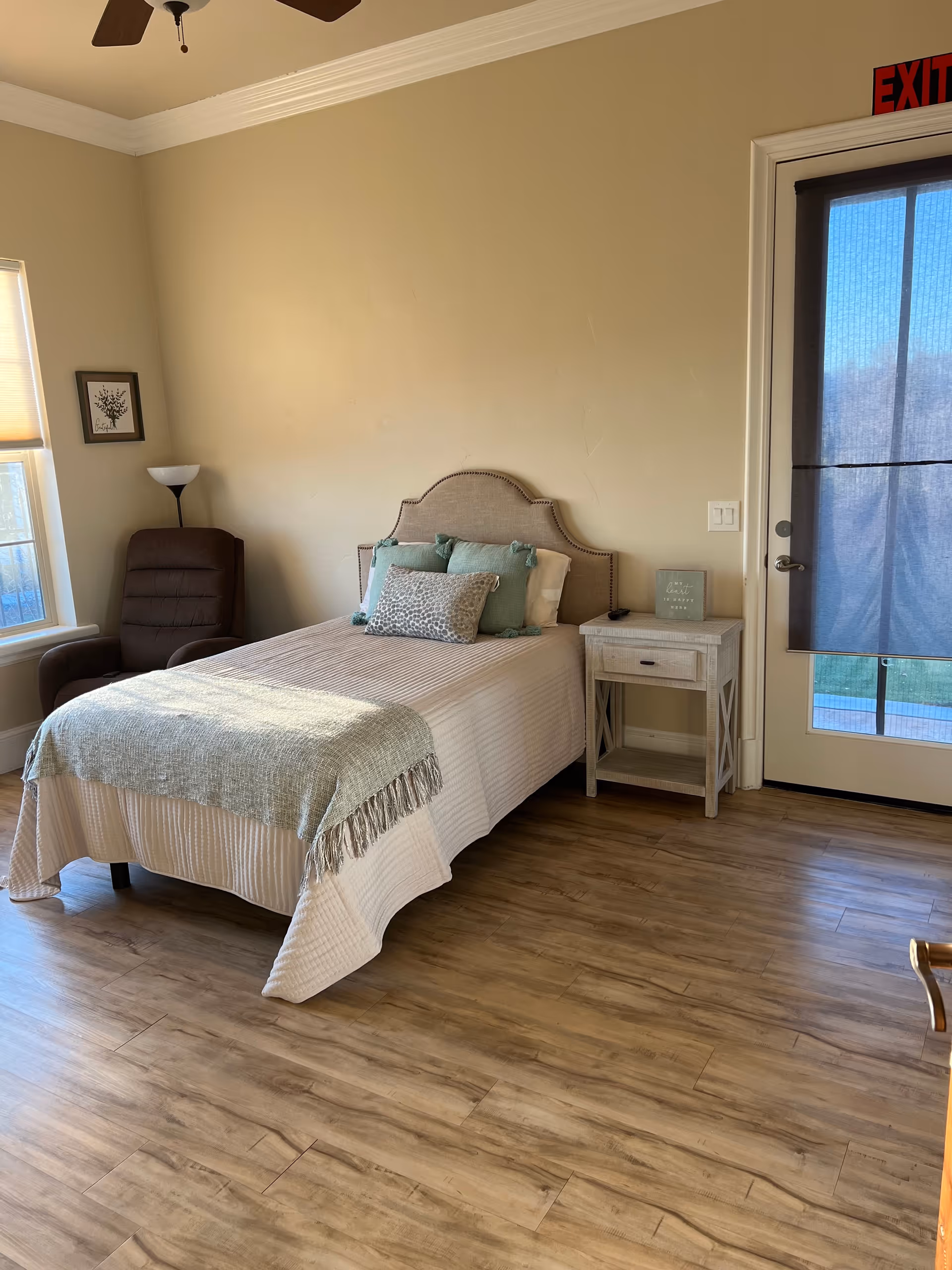 Bright, neatly made single bed in a bedroom with a nightstand, armchair, and a glass door with a shade.