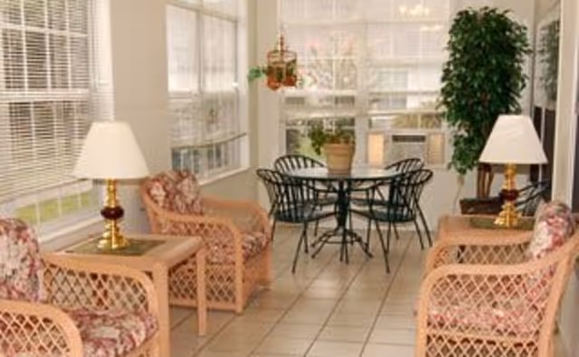 A bright sunroom with tiled floor featuring four wicker armchairs with floral cushions, two side tables each with a lamp, a round glass-top table with four black metal chairs, and a large potted plant near the window.