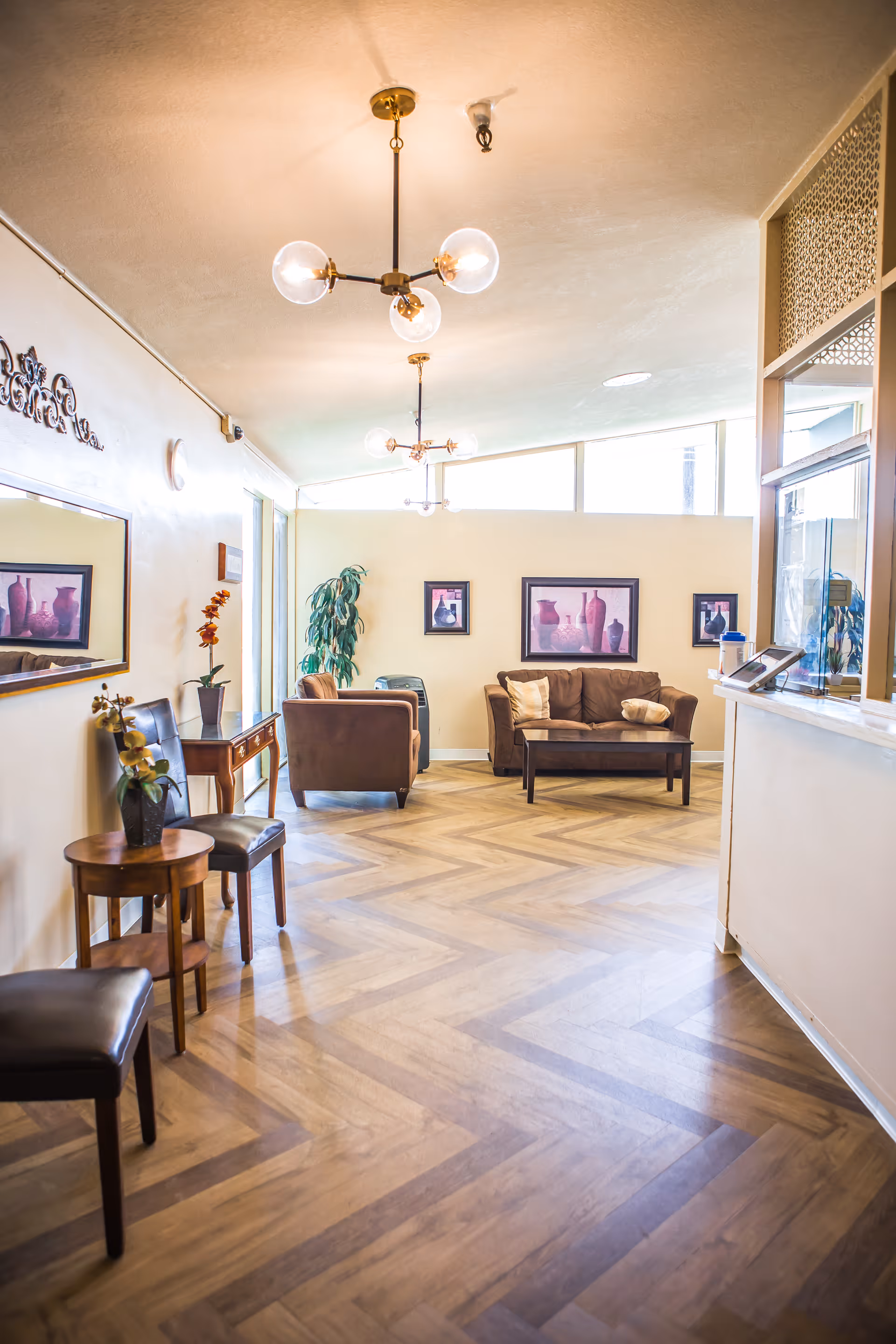 A bright and welcoming interior common area with wooden herringbone flooring, modern ceiling light fixtures, and beige walls. The room features a brown sofa with cushions, a matching armchair, a small wooden coffee table, and several chairs along the wall. There are framed pictures of vases on the wall and a potted plant near the window. A reception desk is visible on the right side of the image.