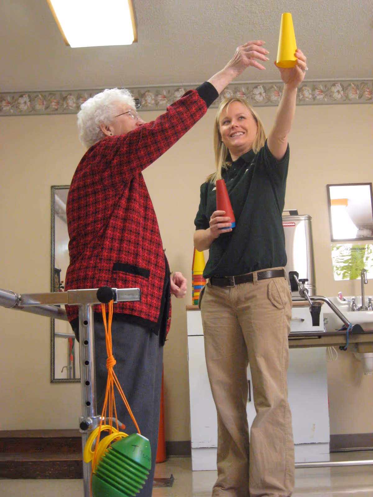 An elderly woman and a younger woman standing indoors, engaging in a physical activity involving stacking or holding colorful plastic cones. The elderly woman is wearing a red and black checkered jacket and using a walker, while the younger woman, dressed in a black shirt and beige pants, is smiling and assisting her.