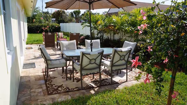 Outdoor patio with a glass table, six cushioned chairs, umbrella, and greenery.