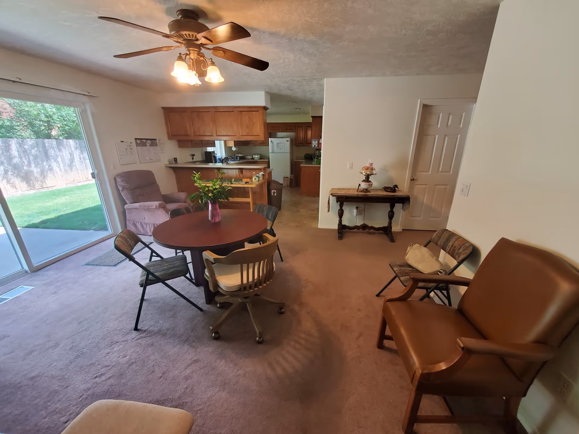 Interior view of a senior living facility room with a round wooden table surrounded by various chairs, a ceiling fan with lights, a sliding glass door leading to a grassy outdoor area, a purple recliner chair, a small wooden table with decorative items, and a kitchen area with wooden cabinets and a refrigerator in the background.