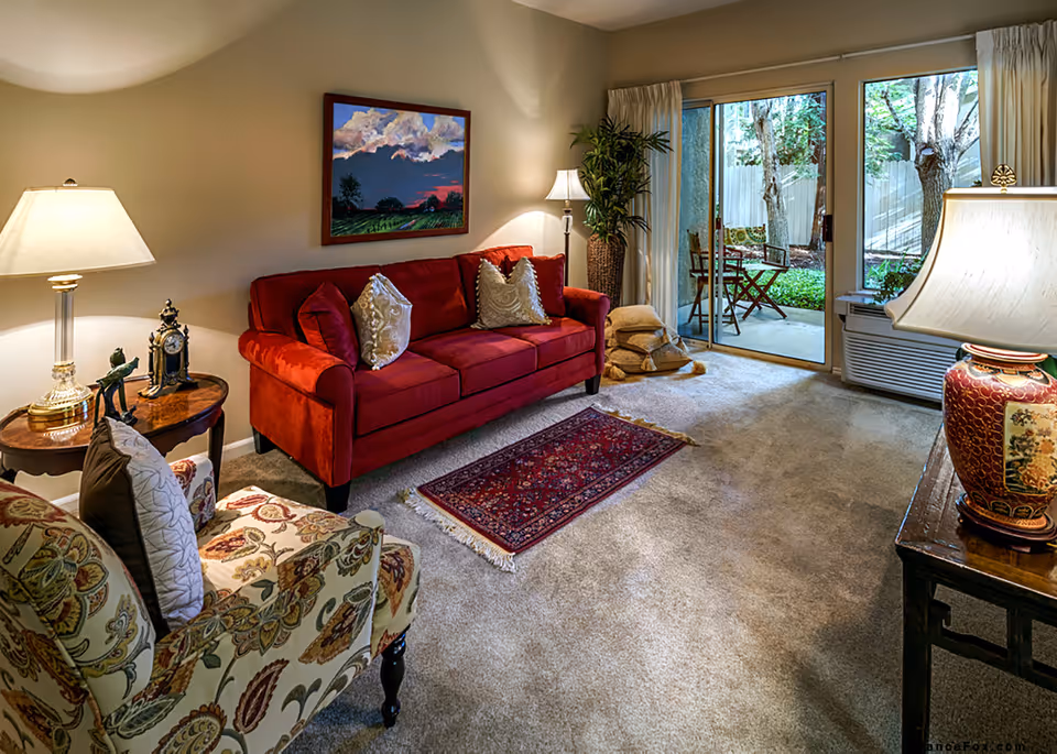Cozy living room with a red sofa, patterned armchair, lamps, rug, and sliding glass doors opening to a small patio.