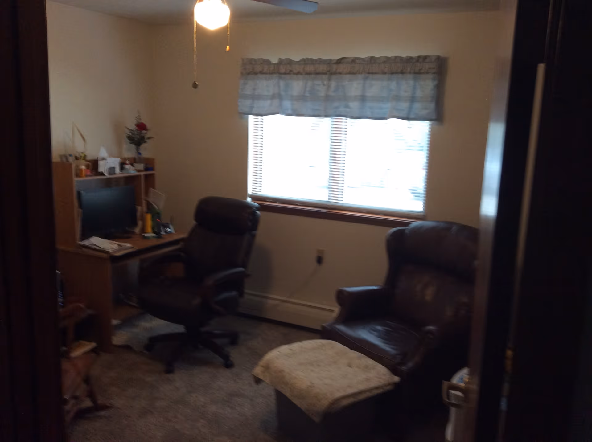 A small room with a window covered by a valance curtain. The room contains a black office chair in front of a wooden desk with a computer monitor and various items on it. There is also a dark leather armchair with a matching ottoman covered by a light-colored cloth. The room has carpeted flooring and a ceiling fan with a light.