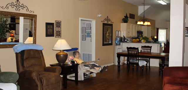 Interior view of a living and dining area in an assisted living cottage. A senior woman is seated in a recliner chair near a side table with a lamp. The room features wooden flooring, a dining table with chairs, a kitchen area in the background, and various wall decorations including framed pictures and a large mirror.