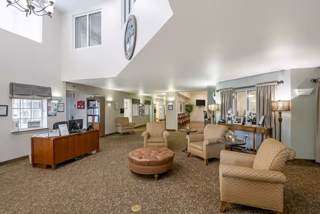 A spacious senior living facility common area with beige patterned carpet, several cushioned armchairs, a round brown ottoman, and a wooden reception desk. The room has high ceilings with a large clock mounted on the wall, windows letting in natural light, and various lamps and framed pictures decorating the space.