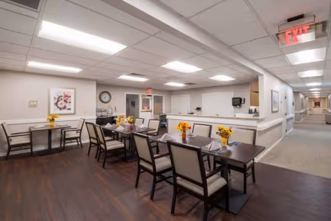A dining area in a senior living facility with several tables and chairs arranged neatly. Each table has a small vase with yellow flowers. The room has a dark wood floor, white walls with a chair rail, and a drop ceiling with fluorescent lighting. There is a hallway visible to the right and an exit sign above a door in the background.